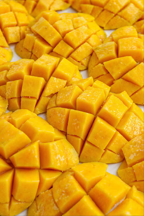 A Mango Sticky Rice candle on a wooden coaster beside a bowl of ripe mango and a plate of sticky rice in warm light